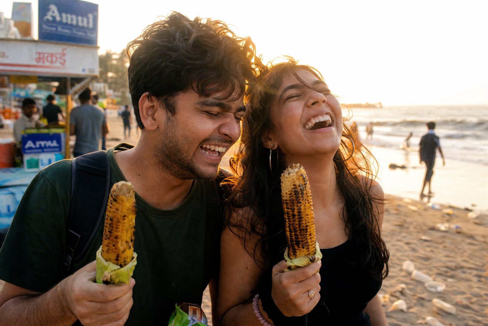 Two young friends laughing over roasted corn at Juhu beach at sunset